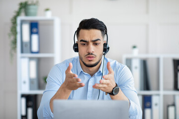 Concerned young Arab man in headset making online video call with business partner, solving problem...