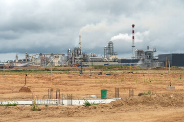 An empty construction site is located next to a large factory. Large factory chimneys emit thick, white, noxious smoke against the gray sky. Environmental disaster and the collapse of civilization.