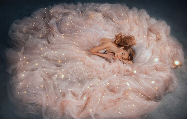 Fairy queen fashion model in luxurious shining pink dress posing in studio. Princess girl, peach outfit with sequins lies in fabrics of outfit, long skirt gown. Fantasy woman sleeping beauty woke up.
