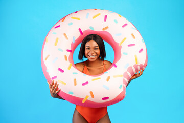 Young black lady in swimsuit posing with donut shaped inflatable ring, smiling at camera over blue...