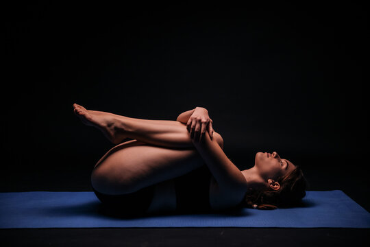 one young woman, exercising yoga, laying on a mat; calmly resting and streching in fetus position. shot on black background.