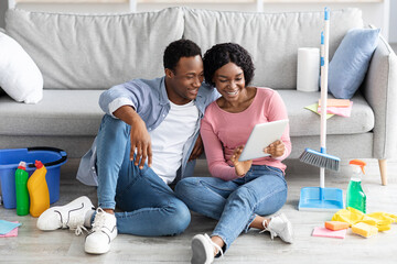 Smiling black couple with cleaning tools resting, using digital tablet