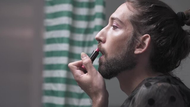 Man painting his lips with red lipstick. Real trans LGBT person with beard primping in the bathroom at home. Diversity Transgender and Gay Concept