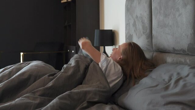Woman in a white T-shirt wakes up happy, stretching, sitting on a comfortable bed, in a modern hotel bedroom