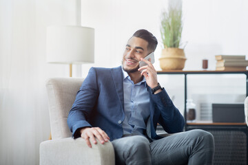 Fototapeta premium Arab businessman talking on mobile phone at office, sitting in armchair, having conversation with friend or client