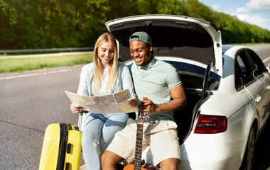 Naklejka premium Multiracial couple going on road trip in car, looking at map, taking break from driving, sitting in auto trunk together