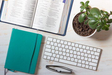 computer keyboard, open bible, notebook and reading glasses and plant on white wood background