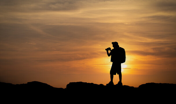 Silhouette of hiker man holding binoculars on sunset from mountain top. Old man with backpack watching the sunset