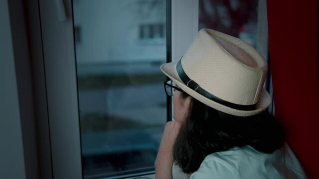 teen girl in a hat looks out the window alone in the evening, cars are passing by on the street, miss, back view