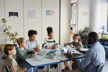 Diverse group of children sitting at table with male teacher in modern school classroom - Powered by Adobe