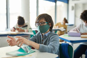 Portrait of young boy sanitizing hands in school classroom, covid safety measures, copy space