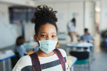 Portrait of teenage African-American girl wearing mask in school classroom and looking at camera,...