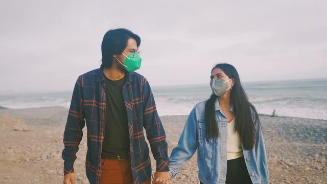 Joven pareja latina de novios enamorados caminando durante un hermoso atardecer en la playa. Personas caminando con mascarillas. Romance en tiempos de pandemia 