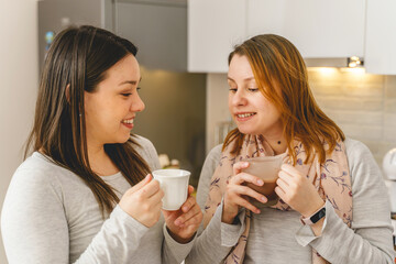 Two women female friends standing at kitchen at home chatting with a cup of coffee or hot chocolate - real people leisure social activity having fun on coffee brake at work