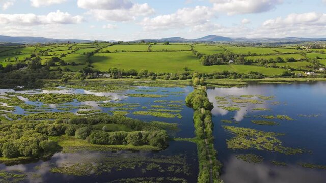 Sunny day with white cloud reflections in the dark blue waters of river Lee and green footpath in the Gearagh, ancient petrified and submerged forest during high tide.