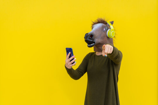 Woman with horse head mask and headphones using smartphone ,pointing finger at camera isolated over yellow background in studio
