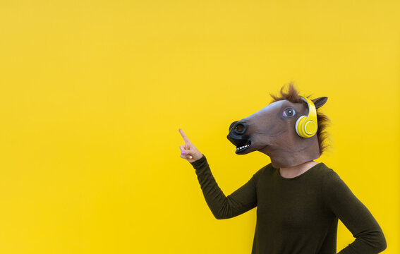 Woman with horse head mask and headphones using smartphone ,points with finger up on blank wall ,indicates copy space isolated on yellow background.