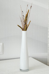 Stylish wooden bedside table and white glass vase with dried branch and plants on it. Interior details. Vertical image