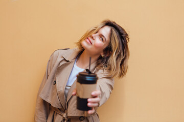 Young smiling millennial woman with wild hair dressed in an autumn coat standing a cup of coffee to go near the beige wall.