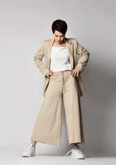Pretty short haired brunette woman in beige business smart casual suit and skeakers standing and posing over light background. Stylish business female wear and fashion concept