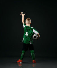 Fototapeta na wymiar Blond smiling boy child in sports green and white clothes standing holding soccer ball and raised hand up over dark background. Trendy sports active children fashion concept