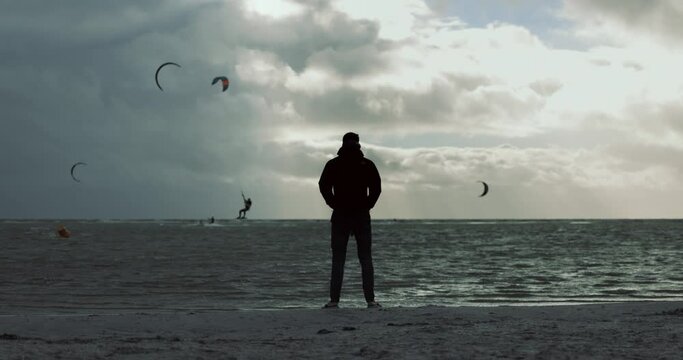 Man Silhouette Standing On Shore Watching Kitesurfers In The Ocean With Dark Clouds Due To Storm Francis. - wide shot