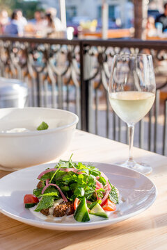 Fresh salad on a table with a glass of white wine at an outdoor cafe, with a wrought iron fence in the background. Bright cheerful image