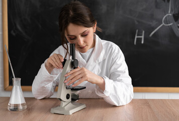 A beautiful woman teacher of biology or chemistry sits at a table against the background of a chalk board. Look through the microscope. White robe. Back to school concept.