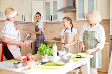 Positive teen girl washes the dishes while others prepare food at table, have fun, talking. focus on female in apron looking at senior grandma