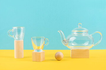 Empty glass transparent tea set. Teapot and and two cups standing on wooden geometric shapes, yellow and blue background. Front view
