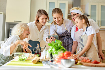 Happy beautiful women use mobile phone while cooking, watching video recipe or tips for cooking, interested in culinary. side view on family dressed in apron, at home in cozy bright kitchen