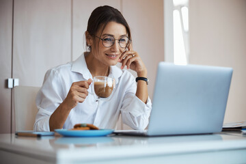 Joyful businesswoman drinking latte and using laptop at work