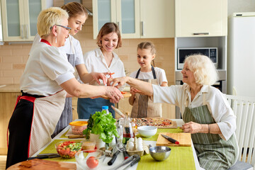 Side view on happy women and kids taking ingredients for pizza, preparing meal, adding missing ingredients, spending good time together at home, wearing apron. portrait of family consisted of females