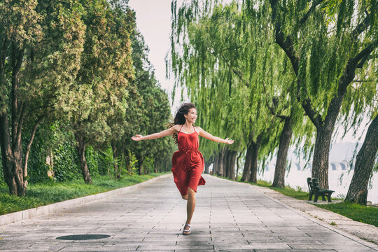 Happy young people summer lifestyle young woman running with open arms in freedom in summer park with trees background. Asian woman in red dress running of happiness.