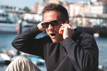 Man with surprised expression talking to his mobile phone at sea port, travel concept