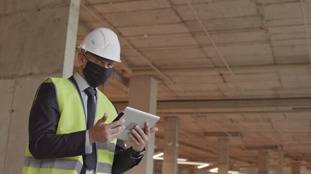 Low angle medium of male Caucasian construction supervisor wearing face mask, green reflective vest and white hard hat, holding tablet computer, then looking on cameraLow angle medium of male Caucasia