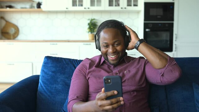 Cheerful African-American man in headphones holding phone and looking at the screen, enjoying watching music video clips, nods head in tact, relaxes and rest sitting on the comfortable couch at home