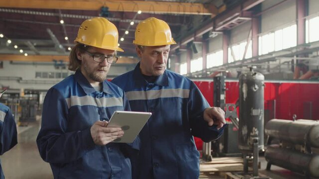 Medium slowmo of senior male supervisor discussing work on plant facility with bearded man holding digital tablet both wearing hard hats and uniforms