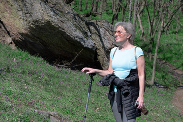 A woman in her 60s stands in the woods and rests from the walk. The concept of an active healthy life. A woman holds a walking stick, a black jacket tied around her waist. Backpack on the back. 