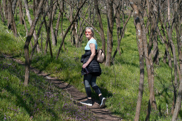 Naklejka na ściany i meble A woman with blonde hair and sunglasses with a bottle of water in her hand, in a sports wardrobe sets through the woods. It is surrounded by bare trees