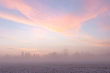 Sunrise over snow covered meadow with ground fog in winter