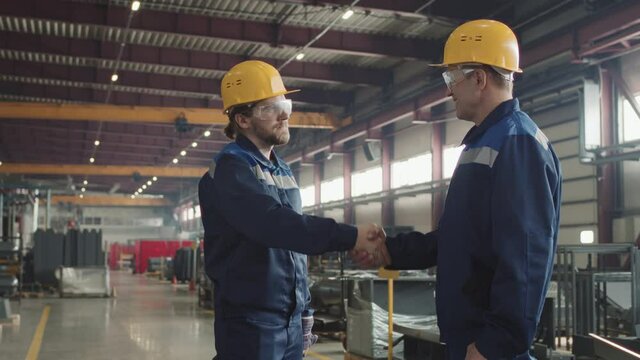 Side-view medium shot with slowmo of two male supervisors in safety glasses and hard hats shaking hands after successful cooperation standing at plant facility