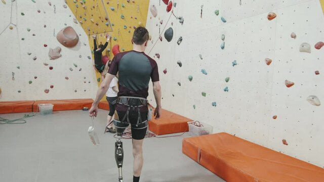 Tracking tilt-up shot of confident athletic man with leg prosthesis walking towards climbing wall in modern sports centre getting ready for climbing