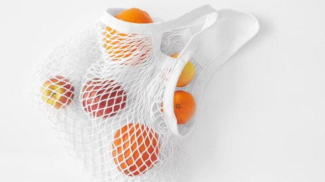 Moving picture of white mesh bag with fruits flat lay on white background top view. Eco friendly, reusable shopping bag. Oranges, apples, garnet in cotton knitted string bag. Zero waste concept.