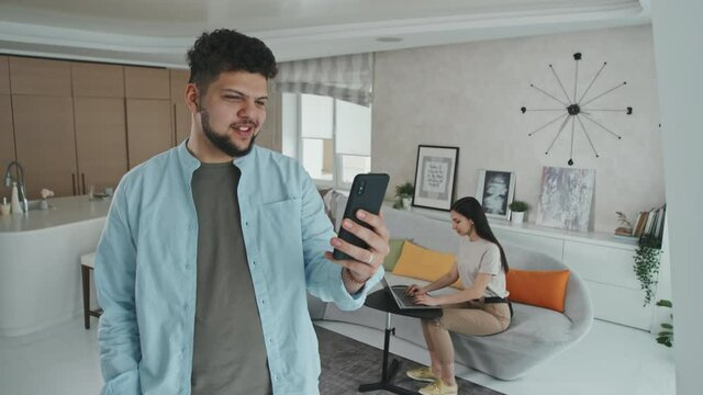 Medium shot of young Latin man video calling with friends or relatives via smartphone while his wife working on laptop sitting on sofa in background in cozy modern living room