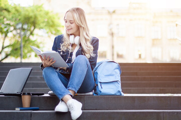Young blond woman is sitting on stairs, studying with laptop computer, books outdoor near college university building. Girl student is preparing for lecture. High school lifestyle. Education concept.