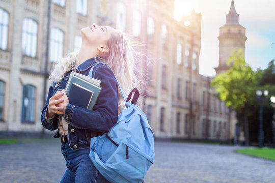 Attractive female student with backpack walking to campus, education concept. Blonde girl is smiling and holding books in her hands on college background. Young woman is taking exams at university.
