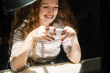 Happy smiling young Caucasian woman drinking coffee while watching video on laptop computer at home in early morning