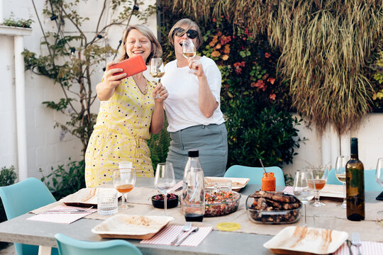 two happy women holding glass of white wine. friends taking selfie with phone at the party. table set with food and drink