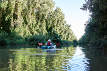 Woman row in blue kayak at wilderness areas of Danube river near green trees and thickets of wild grapes at spring